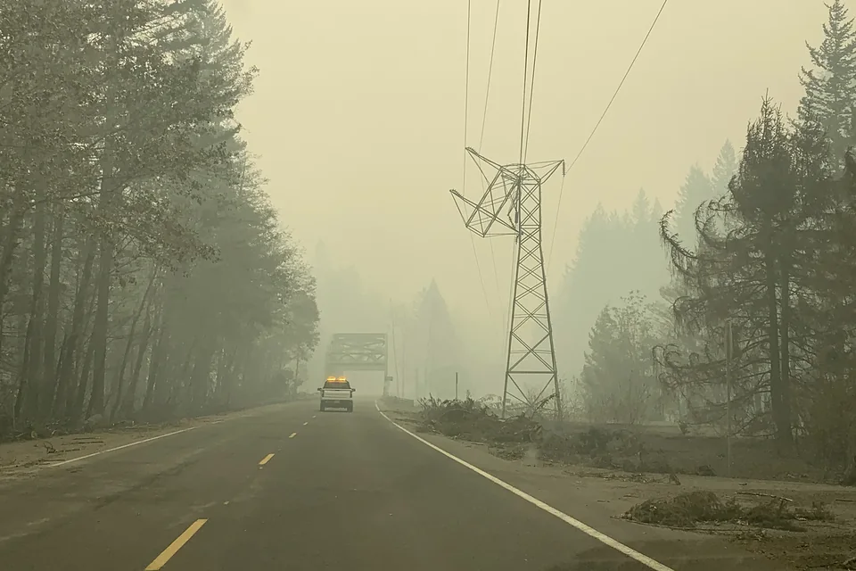 Una torre de transmisión de celosía junto a un camino en un corredor forestal cubierto de denso humo amarillo de incendio